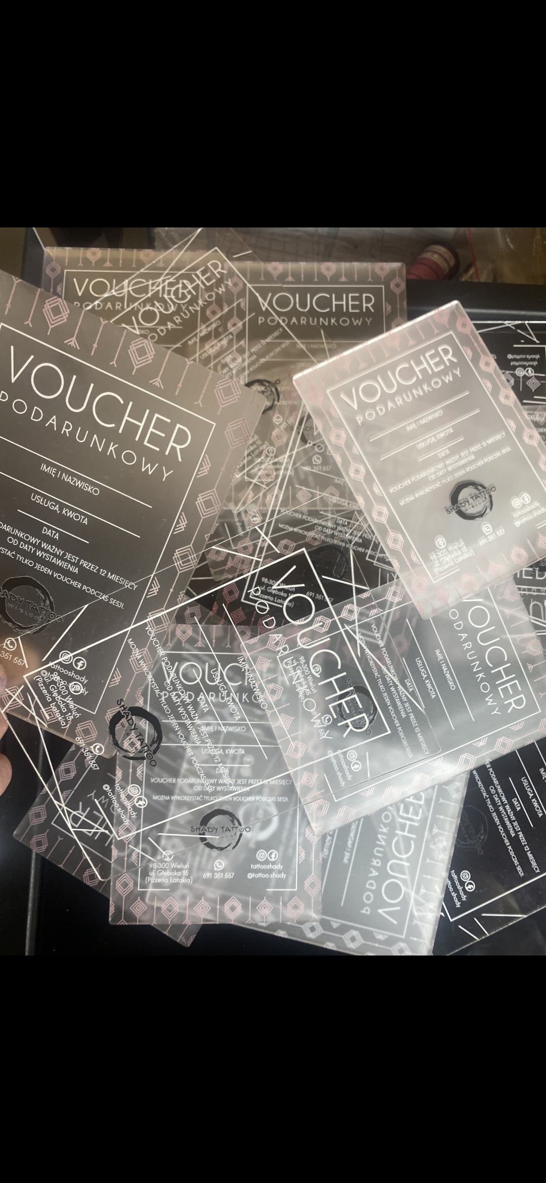 Przezroczysty voucher podarunkowy z pleksi SHADY TATTOO trzymany w dłoni na tle studia przy ul. Głębokiej 15/3 w Wieluniu