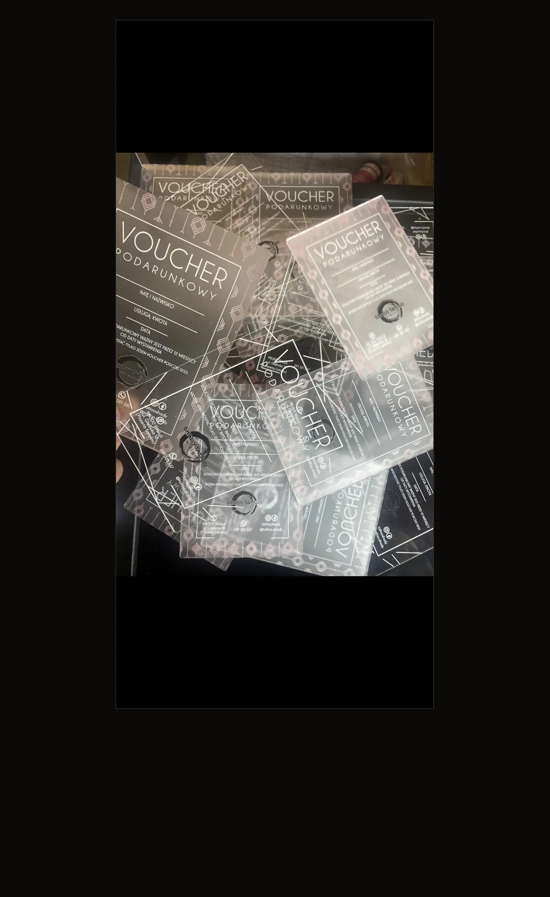 Stos przezroczystych voucherów podarunkowych SHADY TATTOO z pleksi z grawerowanym napisem „Voucher podarunkowy”, logo studia i danymi kontaktowymi, na ciemnym tle
